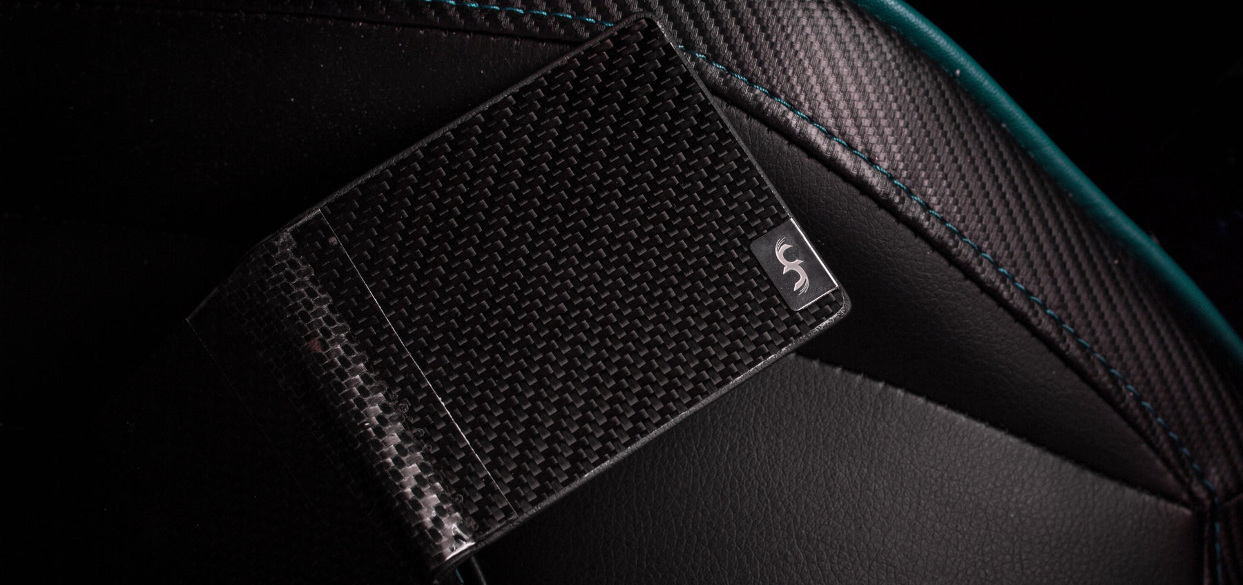 A hinged carbon fiber wallet from Common Fibers sitting on a black and teal interior of a car.